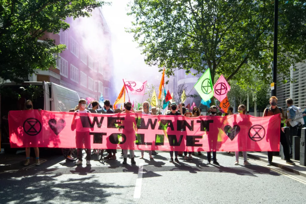 A group of demonstrators carrying a banner reading "we want to live" amidst a peaceful protest, with a colorful smoky background suggesting a vibrant atmosphere.