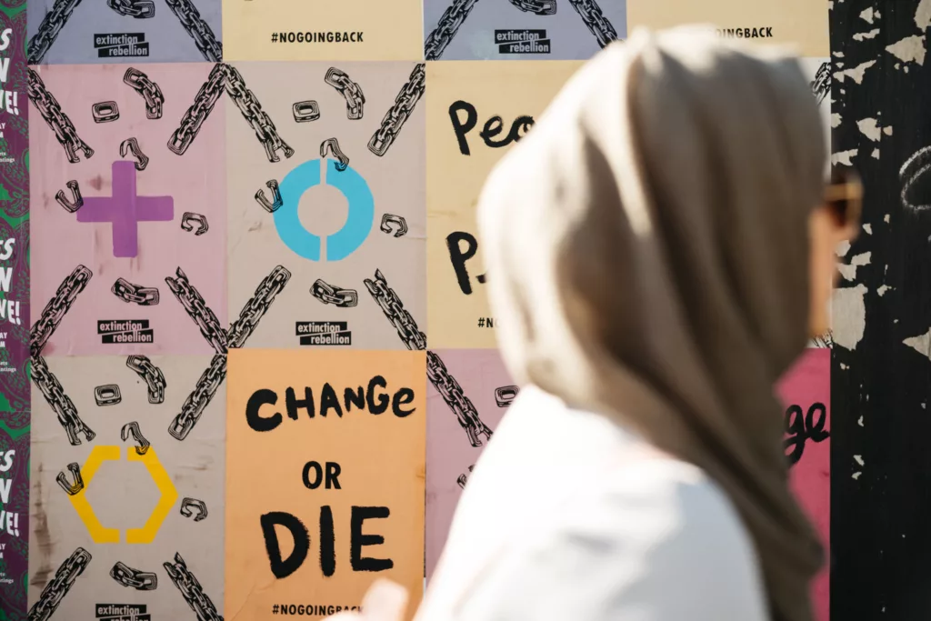 A person in a headscarf observes a colorful wall with bold protest messages and symbols, conveying a sense of reflection on social and political issues.
