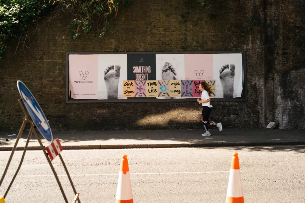 A person jogs past a wall adorned with graffiti-covered posters under the shade of a tree-lined street, while a folding chair and traffic cones occupy the foreground.