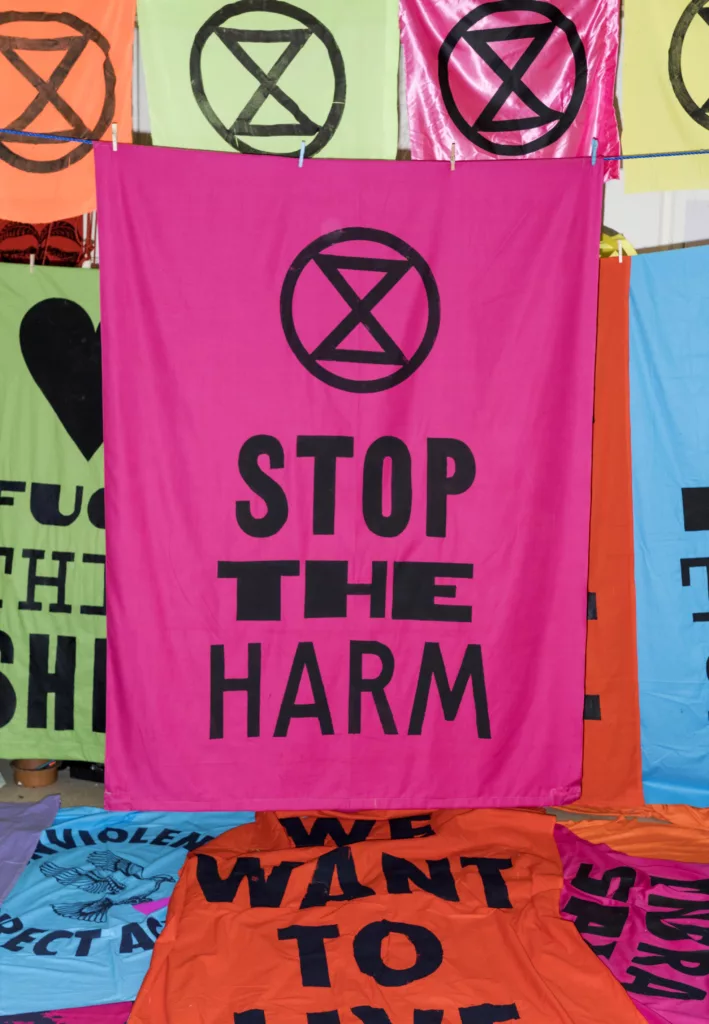 Colorful protest banners with messages advocating for harm prevention, featuring a prominent pink banner reading "stop the harm" with a crossed-out circle symbol above the text.