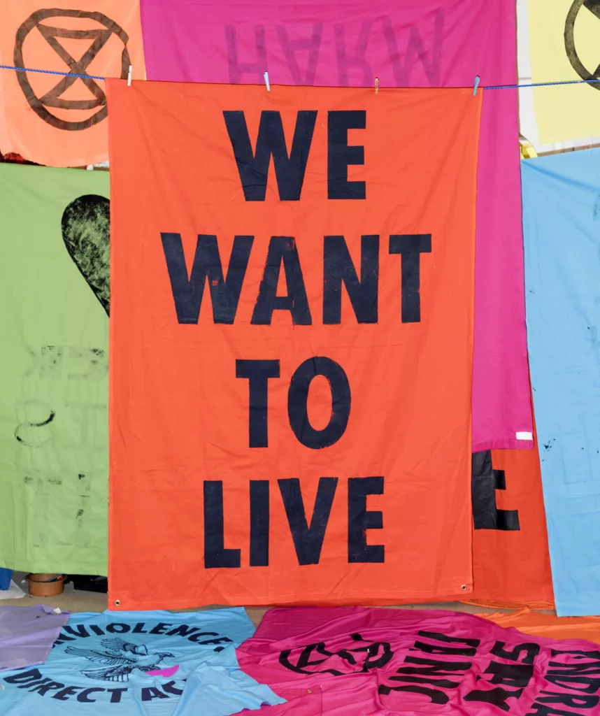 A vivid orange protest banner with bold black letters saying "we want to live" hangs amidst other colorful banners with various messages, symbolizing a call for action and the urgency of social or environmental issues.