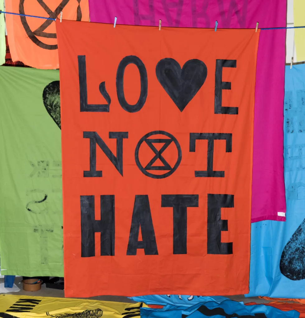 A vibrant red banner with the message "love not hate" in bold black letters, featuring a heart symbol replacing the 'o' in "love" and a peace symbol replacing the 'o' in "not," displayed among other colorful fabrics.