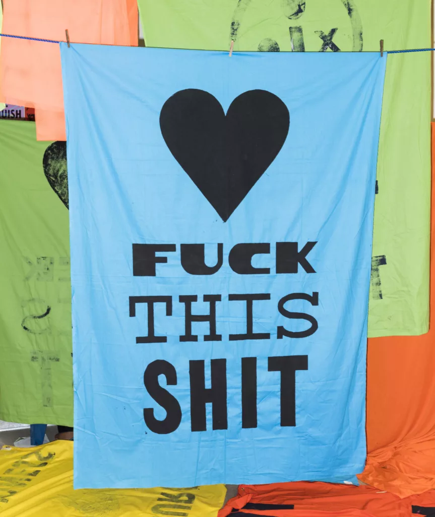 A bold statement banner with a black heart symbol above the phrase "f*** this sh**" hanging on a clothesline against a backdrop of colorful fabrics.