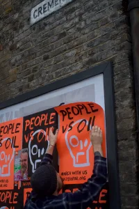 A person is seen putting up a bright orange protest poster with the words "people vs oil" on a public wall, signaling active engagement in environmental activism.
