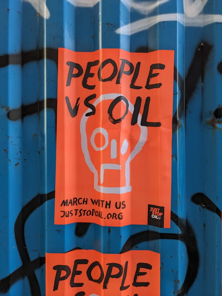 A protest flyer with bold lettering stating "people vs oil" attached to a blue corrugated surface, encouraging public demonstration with a website link for more information.