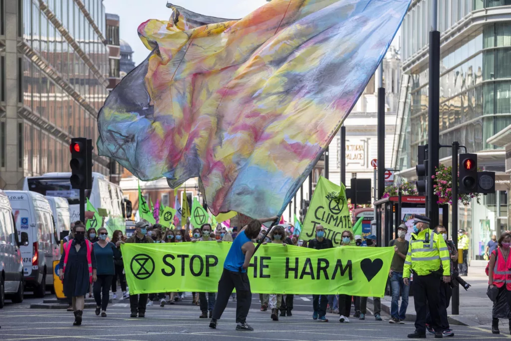Protesters marching with a large colorful banner and a sign reading "stop the harm" during a climate change demonstration in an urban setting, with police officers in the vicinity for crowd control.