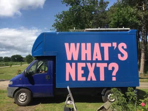 A blue truck parked outdoors with the phrase "what's next?" written in large pink letters on its side.