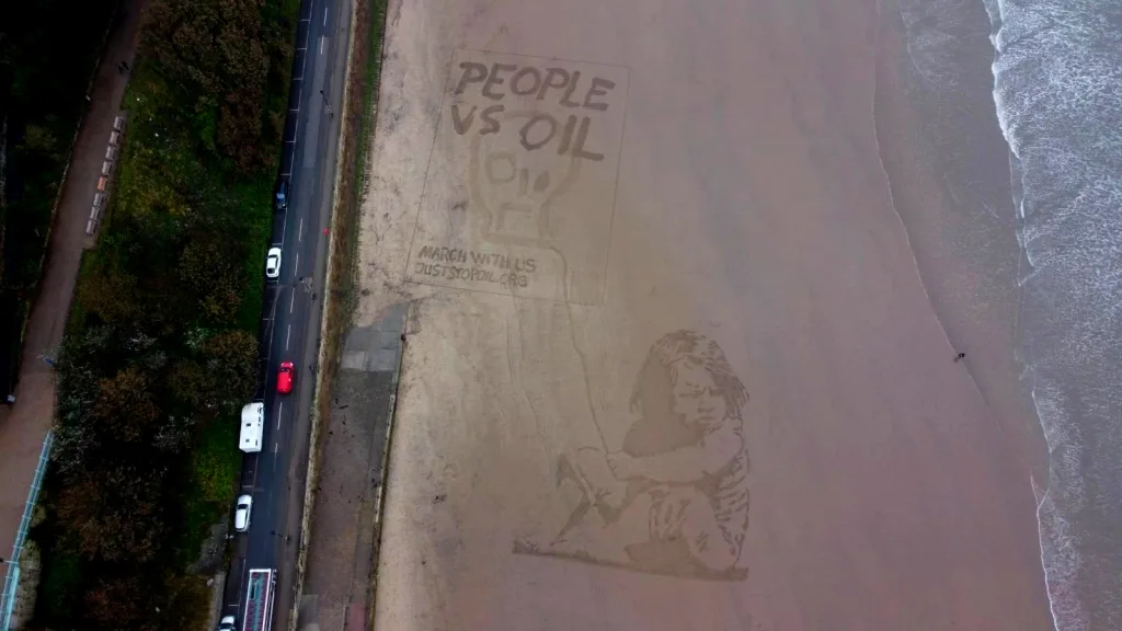 An aerial view of a large environmental-themed sand drawing next to a roadway, featuring the text "people vs oil" and an illustration of a child, with an emphasis on raising awareness about climate change and fossil fuel dependency.