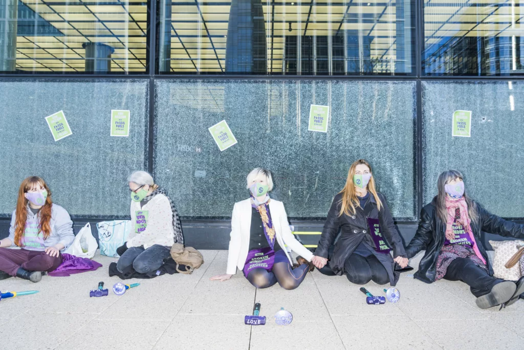 A group of activists engaged in a peaceful protest, wearing face masks and sashes, sitting in front of a building with windows splattered with green paint, symbolizing their commitment to environmental causes.
