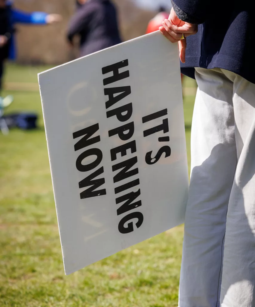 A person holding a sign with the message "it's happening now" at an outdoor event, creating a sense of immediacy and action.