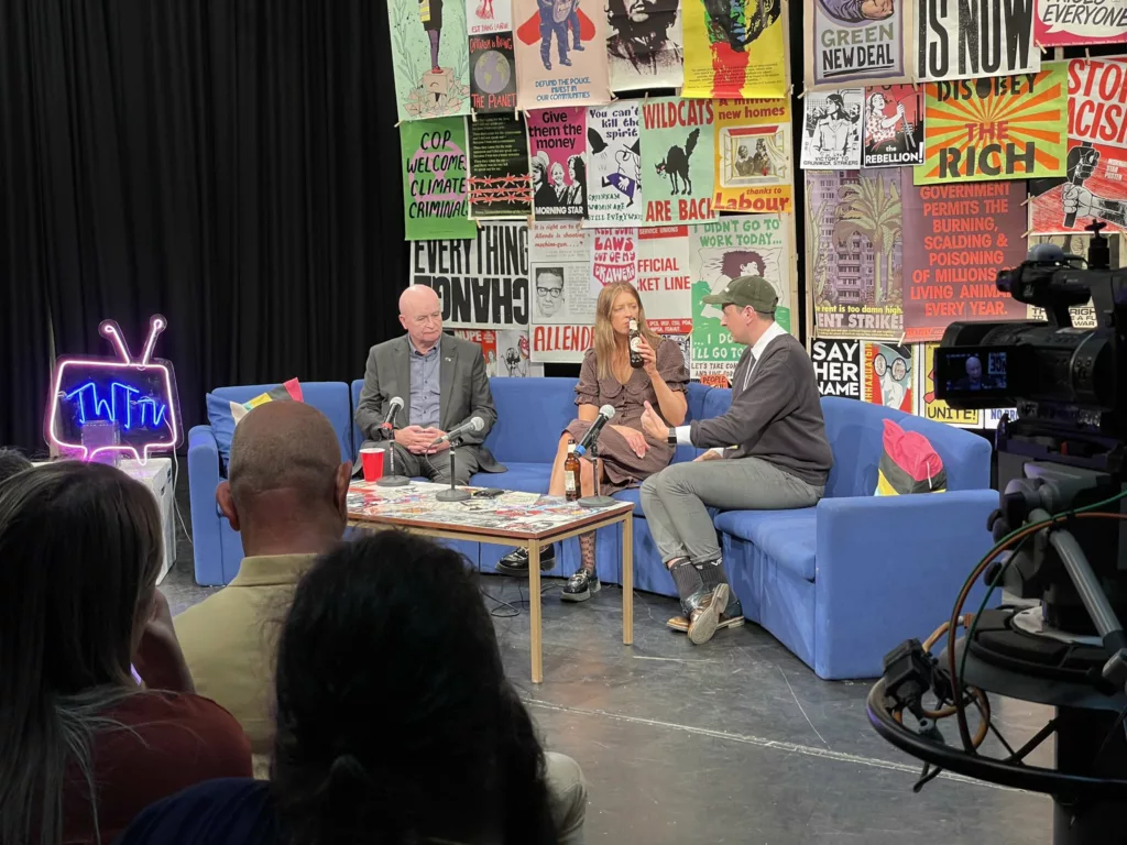 Three individuals engaged in a discussion on a casual studio set, surrounded by vibrant and eclectic wall art, with cameras capturing the conversation for a broadcast or recording.