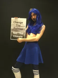 A person in a blue cheerleading outfit holds a placard that says change the politics not the climate