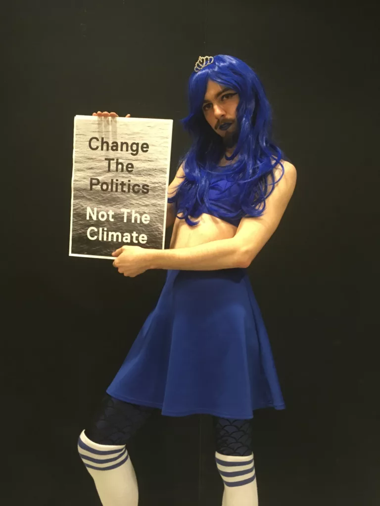 A person in a blue cheerleading outfit holds a placard that says change the politics not the climate