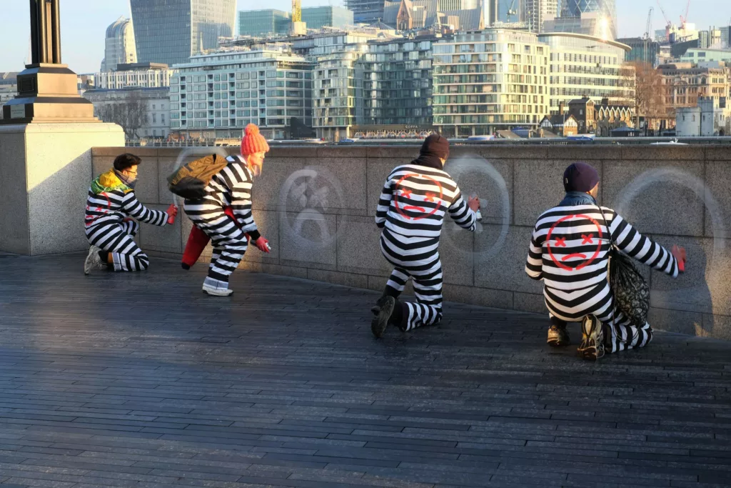 People crouch in black and white stripe suits spray paint unhappy faces on the wall.