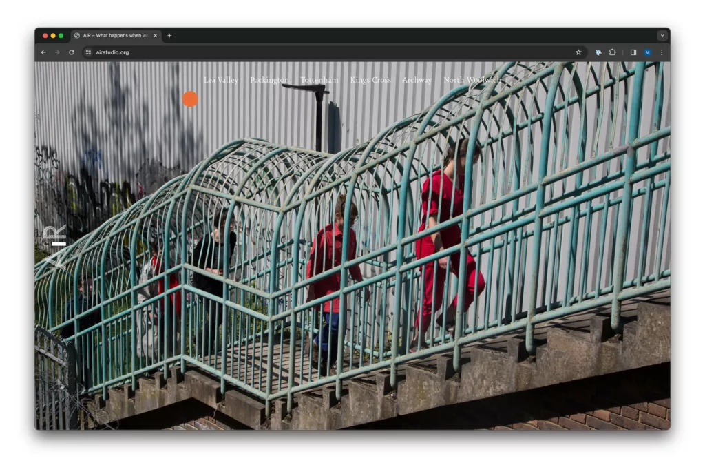 A person in a red jacket walking across a curved green metal bridge with a caged railing on a sunny day.