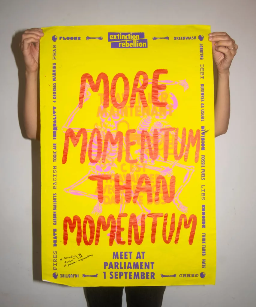 An individual holding up a bold yellow protest sign with the words "more momentum than you momentum" in large red print, featuring various other texts and logos related to environmental activism, suggesting a rally for climate action.