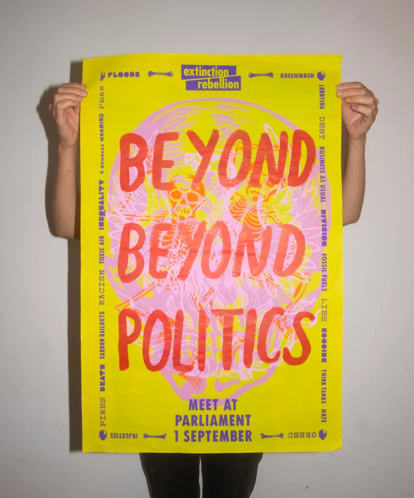 A person holds up a vibrant yellow protest poster related to extinction rebellion with the words "beyond politics" prominently displayed, calling for a gathering at parliament.
