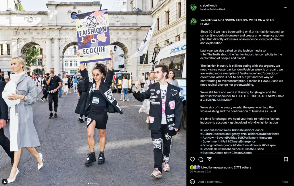 A group of individuals partake in a demonstration for environmental awareness, specifically addressing the fashion industry's impact on the planet, as they march with signs, banners, and a creative display of clothing suspended in the air; against the backdrop of an urban setting with classical architecture.