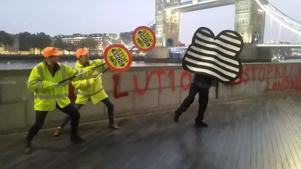 2 lollypop ladies chase someone dressed as a smog cloud. Their sighns say stop smog.